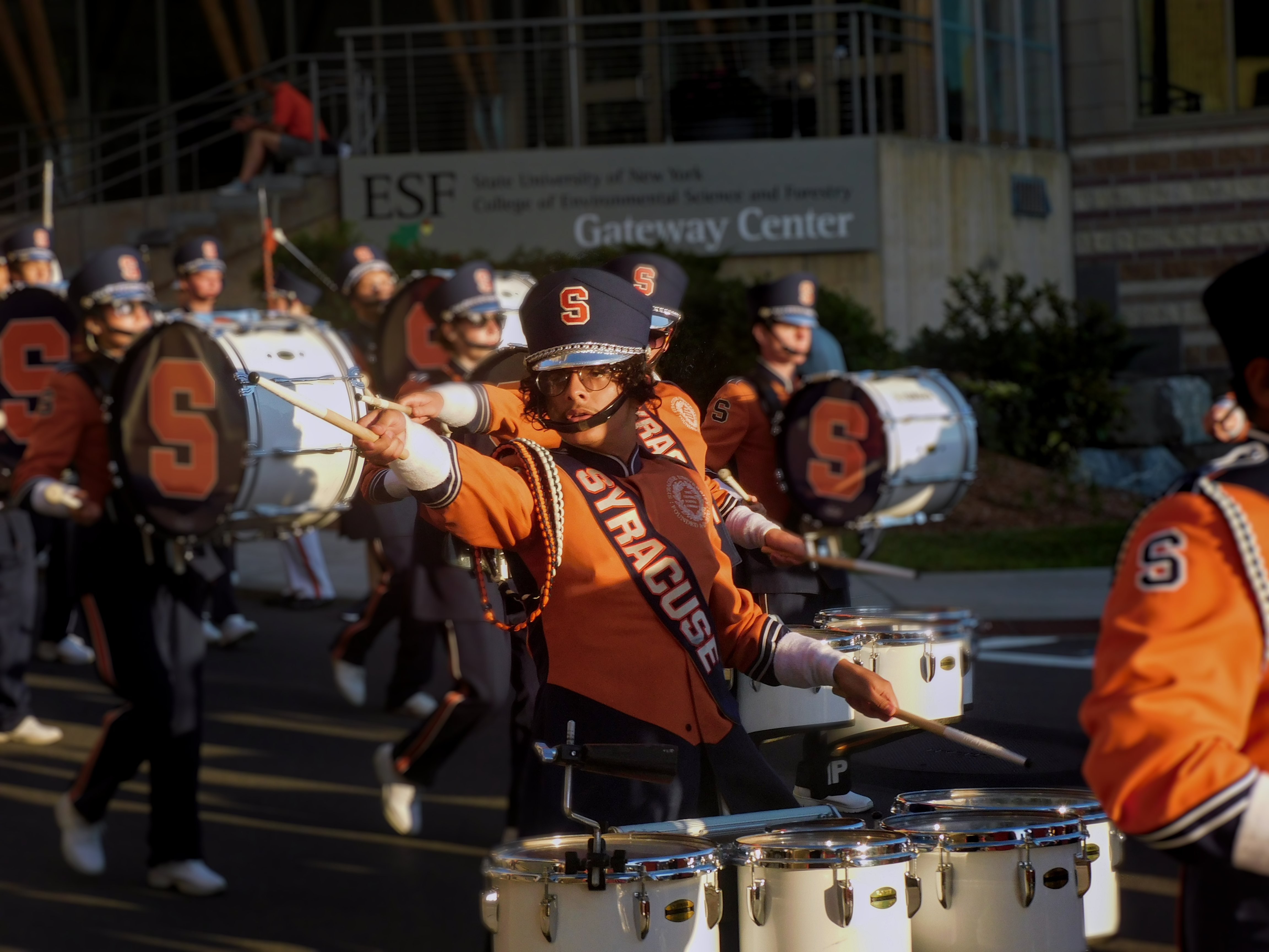 Marching band member in Syracuse University uniform carrying tenor drums and pointing a drum stick towards the left